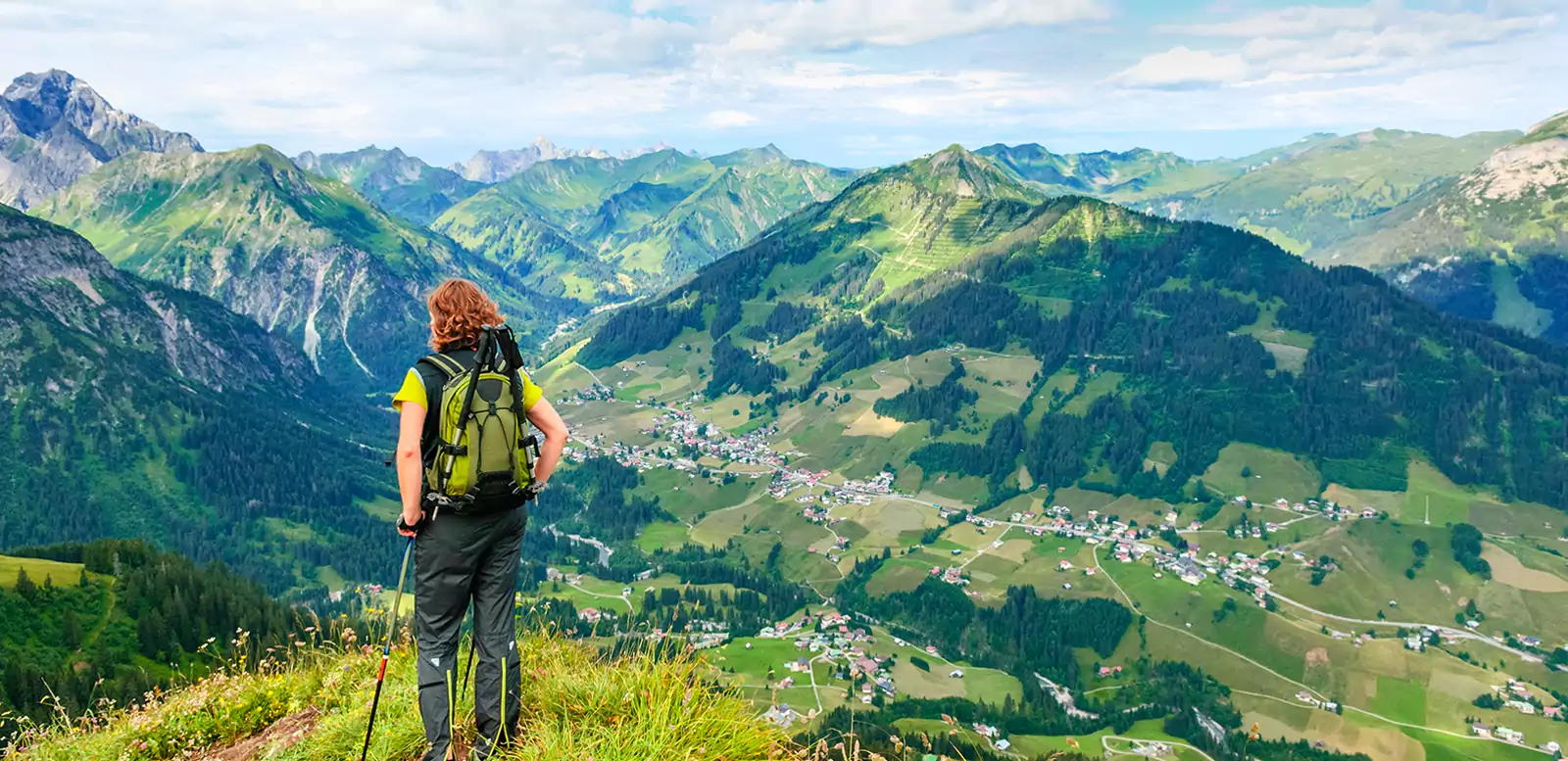 Eine rothaarige Frau in Wanderkleidung steht auf einem Berg und blickt in eine saftig, grüne Berglandschaft