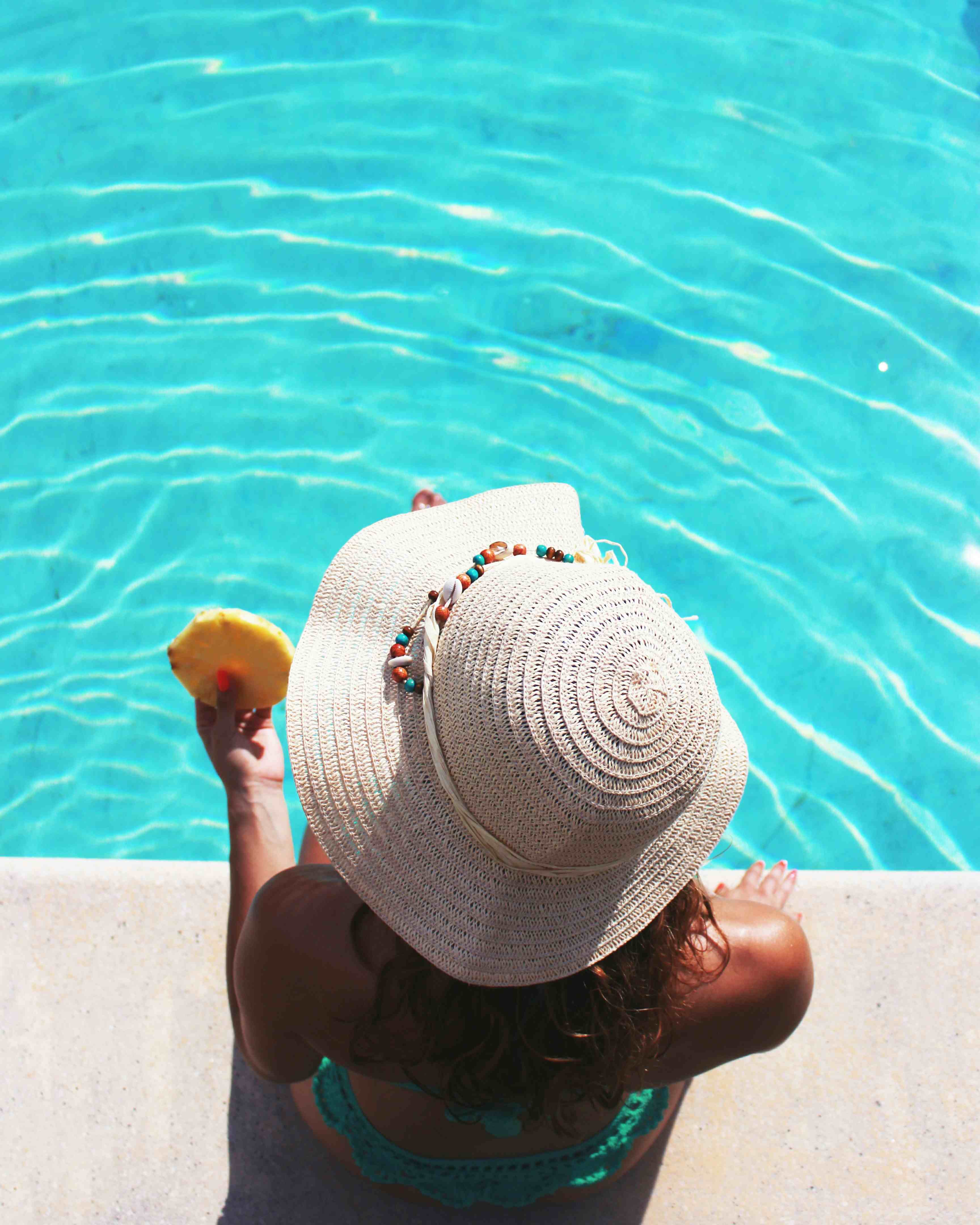 Eine Frau mit Sonnenhut und Ananas in der Hand sitzt am Poolrand