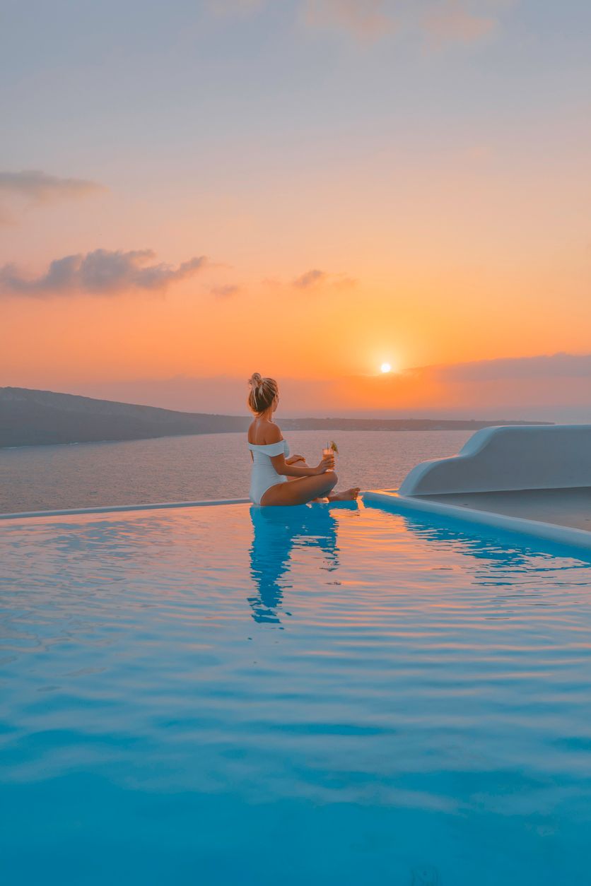 Eine Frau sitzt mit einem Cocktail in der Hand am Rand eines Infinitypools und blickt in den Sonnenuntergang