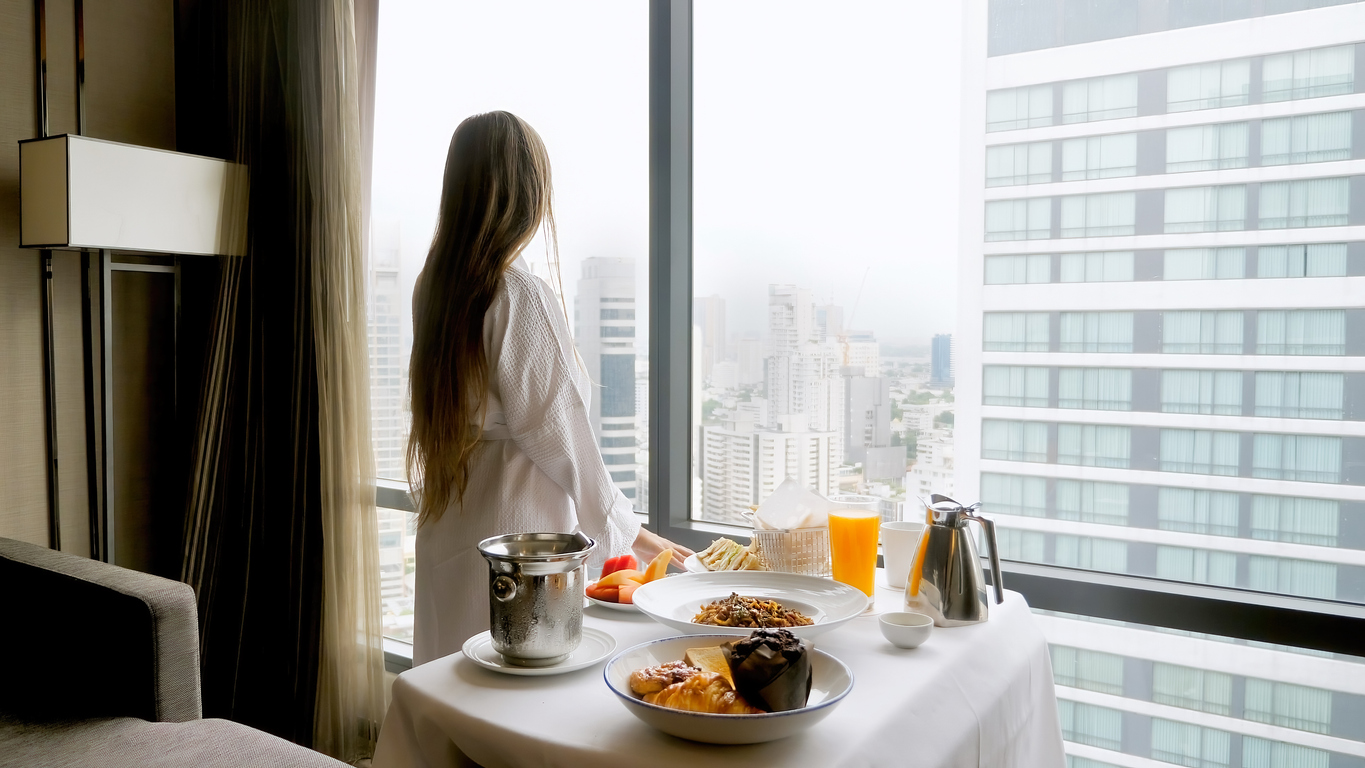 Eine Frau steht im Bademantel an einem gedeckten Frühstückstisch und blickt aus dem Fenster