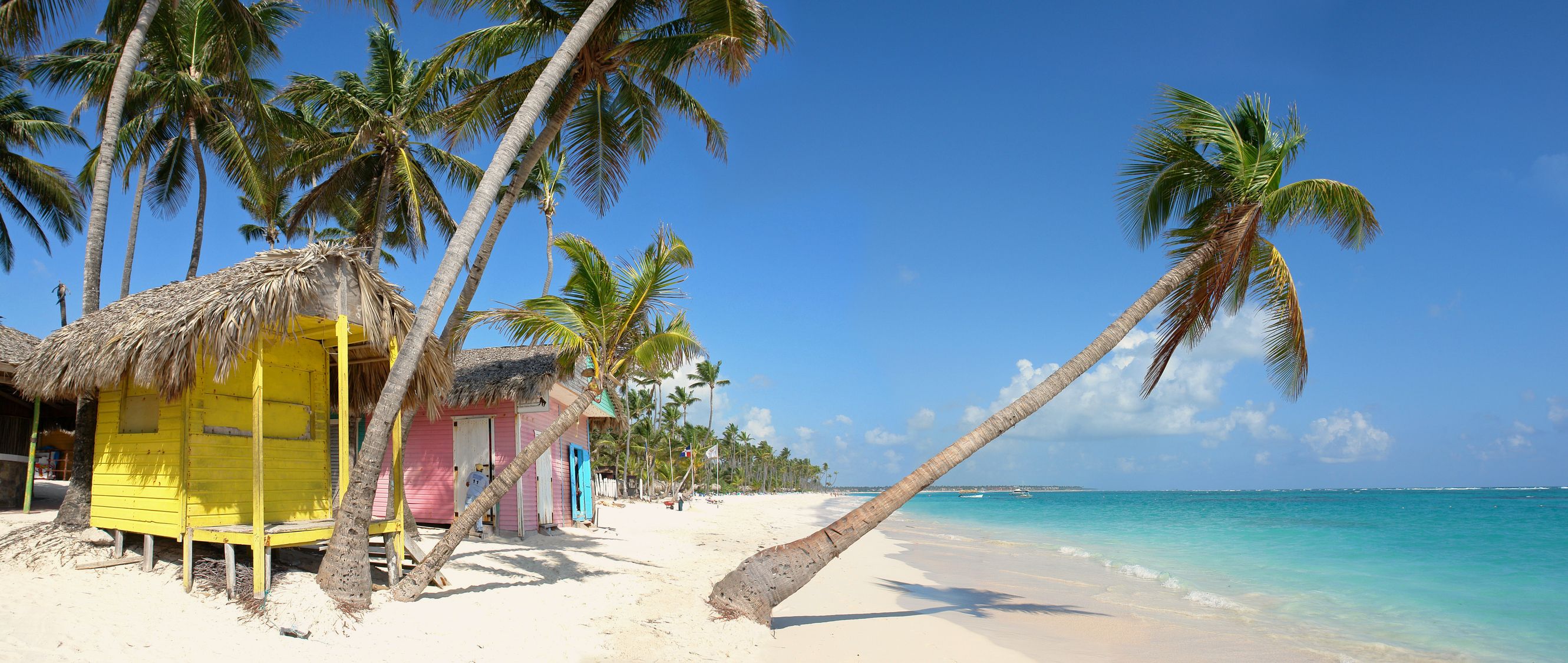 Ein weißer Strand mit bunten Holzhäusern und schiefen Palmen