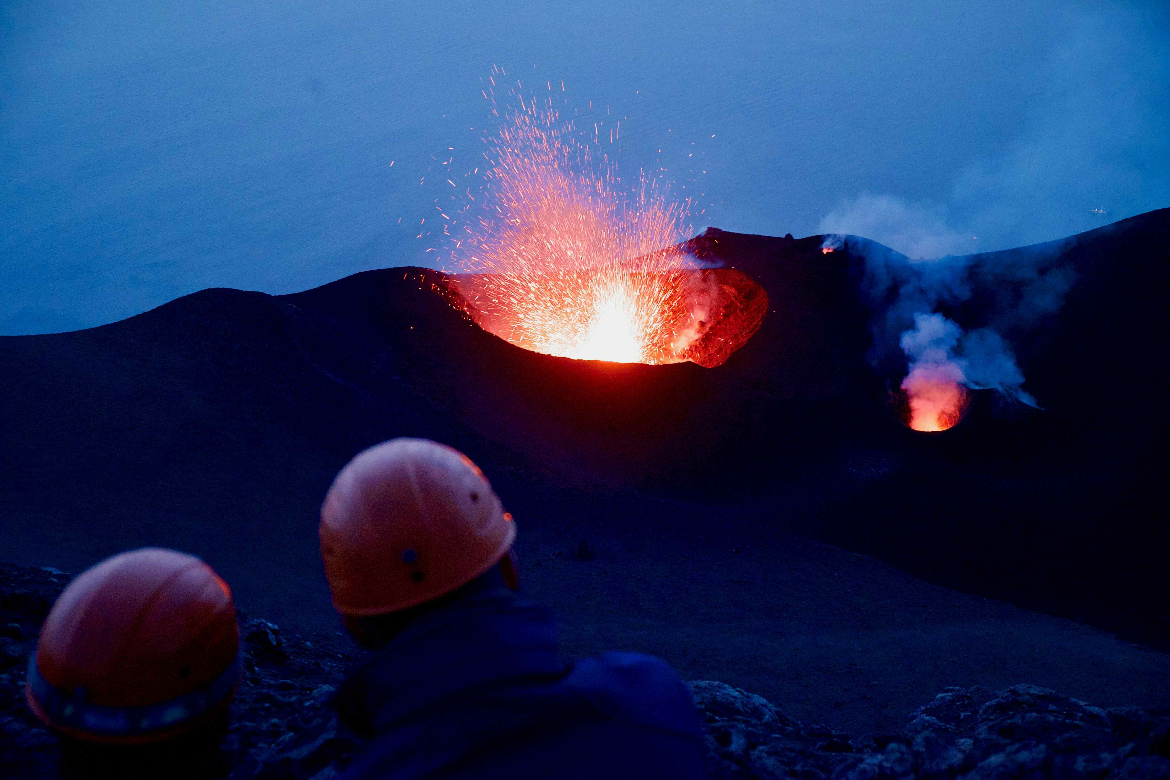Aus einem aktiven Vulkan wird heiße Lava geschossen und vorne stehen zwei Menschen mit Helmen