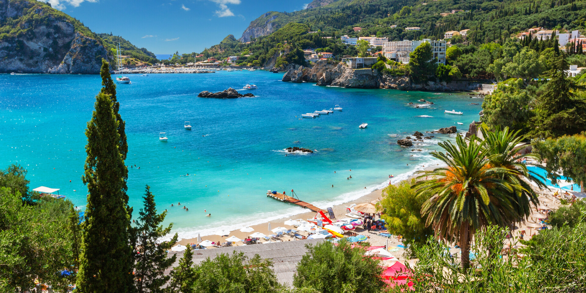 Ein belebter Strandabschnitt zwischen Palmen, grünen Bergen und Häfen