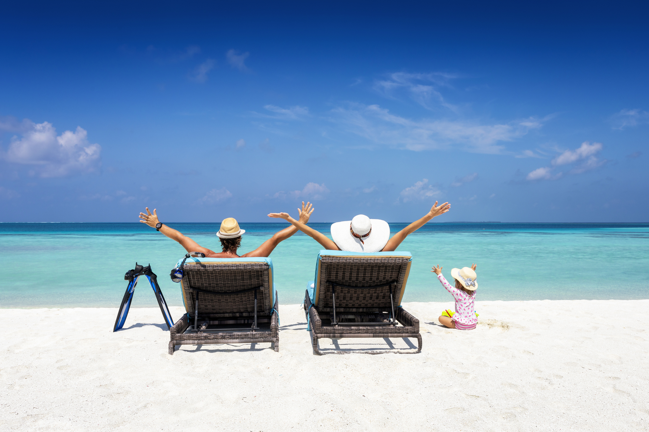 Familie mit Kleinkind sitzt am Strand und guckt auf das azurblaue Meer