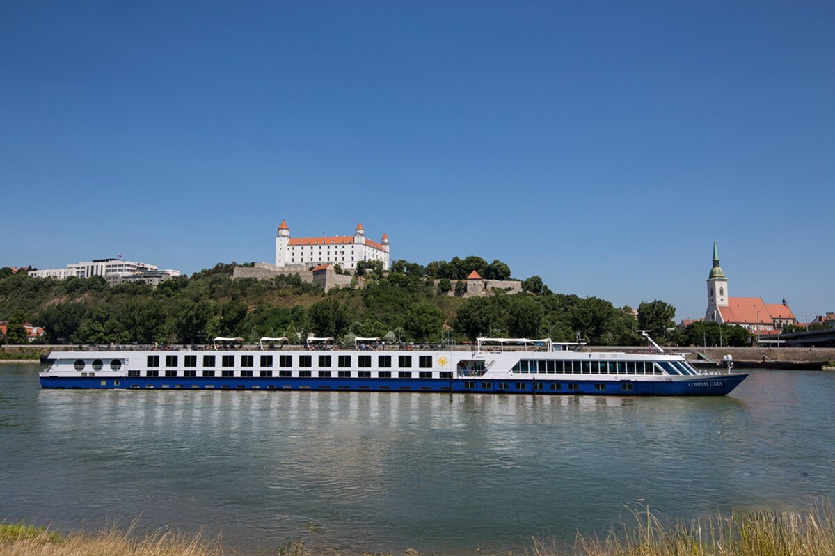 Ein langes, weiß-blaues Flusskreuzfahrtschiff der Reederei Compass Cara gleitet auf einem glatten Fluss an einer Stadt vorbei