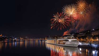 Ein Flusskreuzfahrtschiff liegt im Dunkeln an einem städtischen Hafen mit Feuerwerk am Nachthimmel