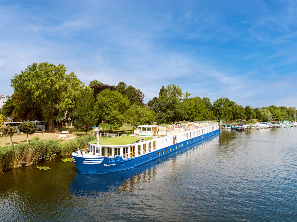 flusskreuzfahrt-thurgau-silvester-elbe Ein weiß-blaues Flusskreuzfahrtschiff der Reederei Thurgau liegt an einem idylischen kleinen Hafen im Grünen an