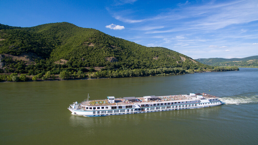 Ein weißes Flusskreuzfahrtschiff der Reederei riverside gleitet durch einen grünen Fluss