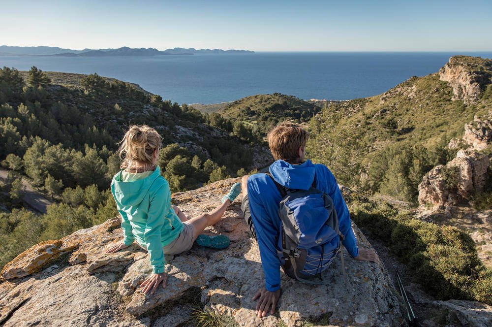 Ein junges Paar sitzt in Wanderkleidung auf einem Felsen und blickt auf das Meer von Mallorca