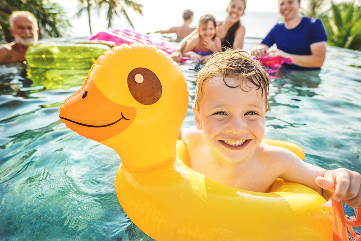 Ein Junge badet in einer gelben Gummiente in einem Pool und grinst in die Kamera. Die Familie ist im Hintergrund.