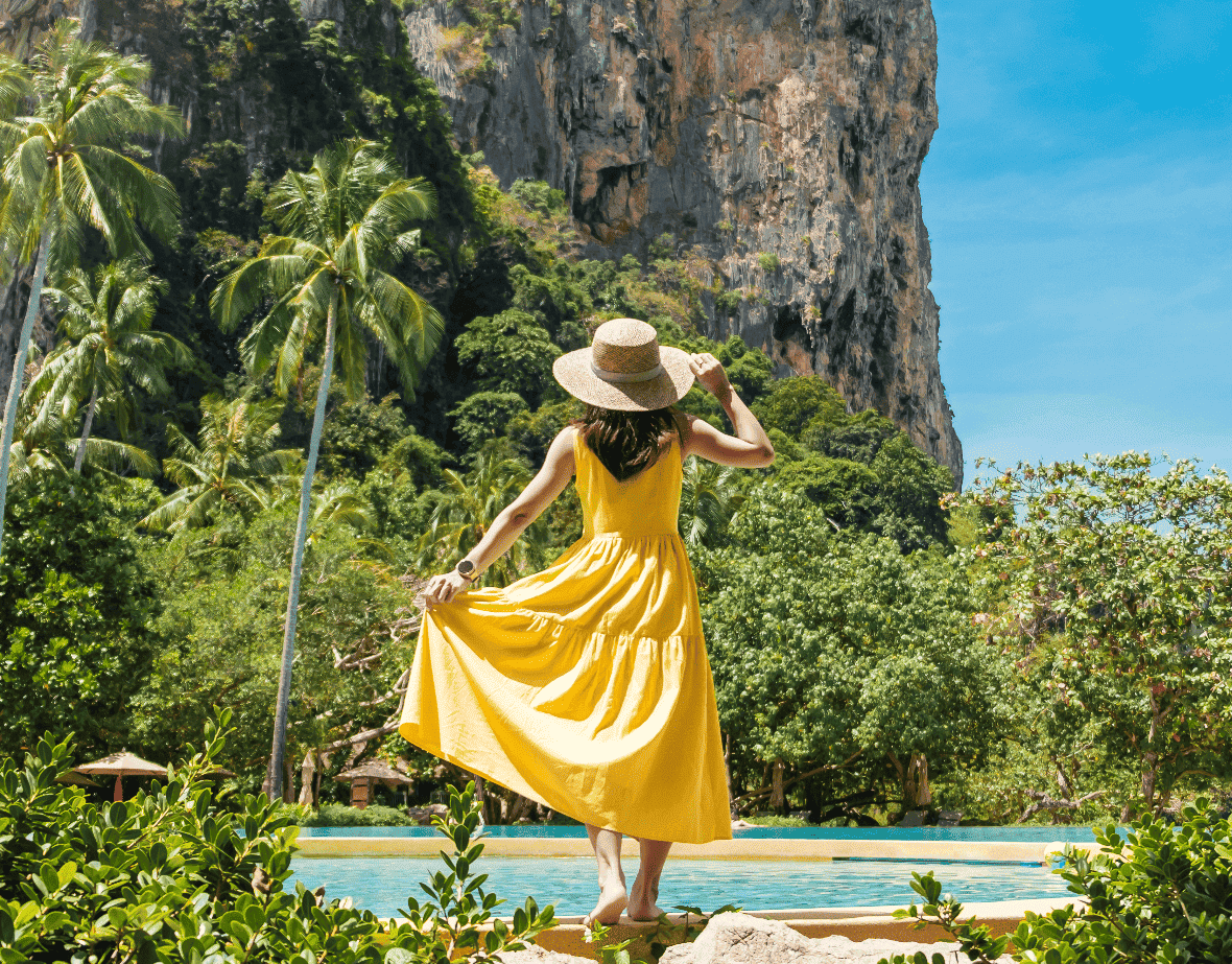 Eine Frau im gelben Sommerkleid und Sonnenhut steht auf einem Felsen. Sie blickt auf ein Panorama aus tropischen Pflanzen, Palmen und gigantischen Kalksteinfelsen am Meeresrand.