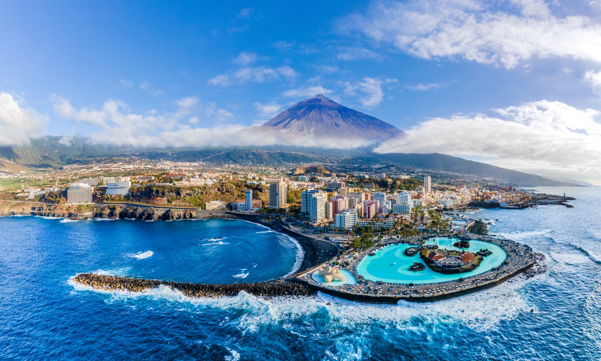 teneriffa-puerto-de-la-cruz-teide Eine Küste mit schwarzem Strand grenzt an eine Stadt mit vielen Hochhäusern, im Hintergrund der Teide